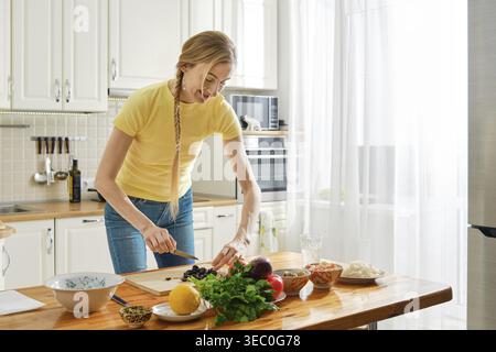 Eine Frau steht in einer Küche und schneidet Oliven auf einem Holztisch. Sie konzentriert sich auf ihre Aufgabe mit verschiedenen Zutaten, die um sie herum platziert werden. Helles Licht kommt Stockfoto