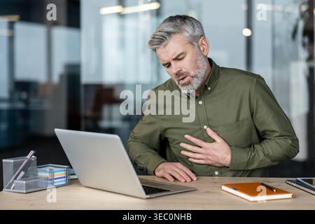 Reifer Mann mit starken Brustschmerzen an seinem Schreibtisch. Er hält seine Brust unangenehm und sieht verstört aus, während er vor einem Laptop sitzt. I Stockfoto