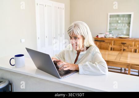 Digitale Inklusion und gesundes Altern, wobei eine ältere Frau zu Hause aktiv einen Laptop benutzt. Sie lächelt, während sie auf der Tastatur schreibt, Showca Stockfoto