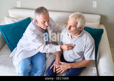 Senior Healthcare — ein männlicher Arzt in einem weißen Mantel und blauen Peelings verwendet ein Stethoskop, um eine medizinische Untersuchung bei einem älteren männlichen Patienten sitzend durchzuführen Stockfoto