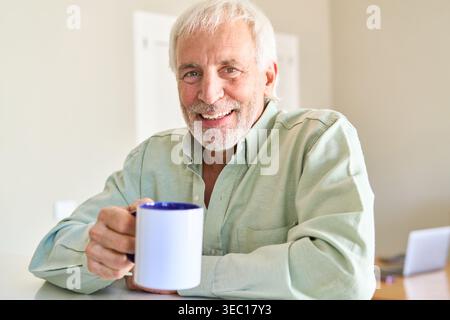 Gesundes Altern und positiver Ruhestand. Ein lächelnder älterer Mann mit grauen Haaren und Bart hält eine Kaffeetasse und blickt direkt in die Kamera Stockfoto