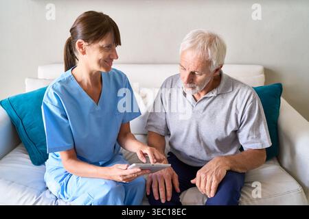 Senior Healthcare Consultation — eine Krankenschwester in blauen Peelings erklärt einem älteren Mann medizinische Informationen mit einem digitalen Tablet. Stockfoto