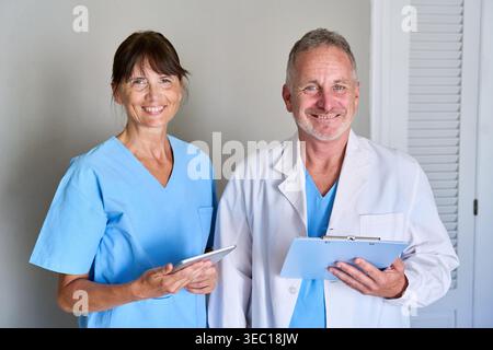 Medizinisches Fachpersonal – ein selbstbewusster männlicher Arzt in einem Labormantel mit einem Klemmbrett und eine Krankenschwester in blauen Peelings mit einer Tablette, beide lächeln an Stockfoto