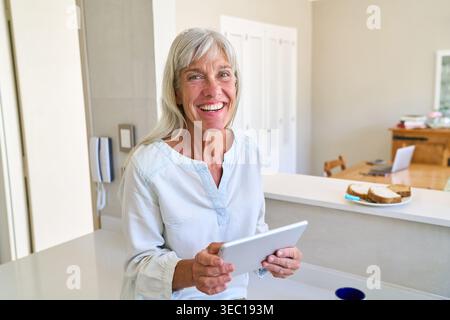 Digitale Inklusion und gesundes Altern – eine lächelnde Seniorin mit grauem Haar hält ein Tablet in ihrer modernen Küche und verkörpert aktiv und unabhängig Stockfoto