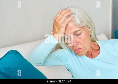 Eine ältere Frau mit Kopfschmerzen oder Migräne, die an Schmerzen und Unwohlsein leidet. Sie wird zu Hause auf einem Sofa gezeigt, mit der Hand auf ihrem f Stockfoto