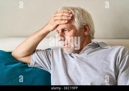 Gesundheitliche Bedenken und gesundheitliche Zustände alternder Erwachsener. Ein älterer Mann mit Kopfschmerzen wird gezeigt, der auf einer Couch ruht und seine Stirn unbehaglich hält. Stockfoto