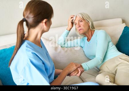 Senior Healthcare Support — eine mitfühlende Krankenschwester in blauen Peelings hält die Hand einer älteren Frau und bietet Komfort und Empathie. Stockfoto