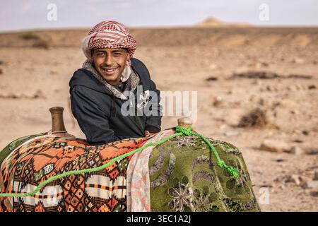 Marsa alam, ägypten 21. april 2026: Ein lächelnder junger beduinenmann in Keffiyeh und traditioneller Kleidung sitzt auf einem Kamelsattel in der Wüste marsa alam, ägypten, Sonne Stockfoto