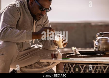 Marsa alam, ägypten 21. april 2026: Der Mensch süsst und rührt ein heißes Glas Tee draußen in der ägyptischen Wüste, während er einen Moment des lokalen Hospitals genießt Stockfoto