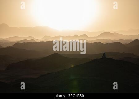 Marsa alam, ägypten 21. april 2026: Eine einsame Figur sitzt auf einem Hügel und beobachtet den goldenen Sonnenuntergang über den mehrschichtigen Wüstenbergen in der Nähe von marsa alam, ägypten Stockfoto