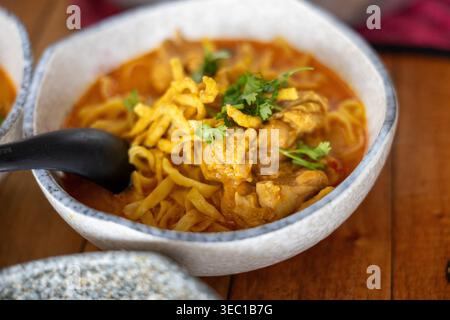Köstliche heiße, scharfe Kokos-Curry-Nudelsuppe mit zartem Huhn und knusprigem Topping in rustikaler Keramikschale auf Holztisch, der sehr gut aussieht Stockfoto