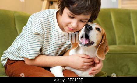 Warmes Nahporträt eines Kindes, das einen Beagle auf einer gemütlichen grünen Couch umschließt, während der Hund fröhlich und entspannt aussieht. Haustiere, Zuneigung und Ruhe drinnen Stockfoto