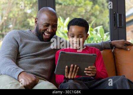 Afroamerikaner mit grauem Pullover und Junge, der auf einer braunen Couch sitzt und lacht Stockfoto