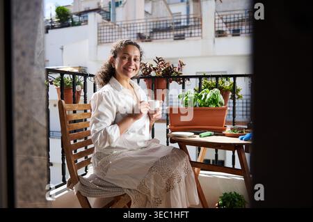 Die lächelnde Frau sitzt auf einem sonnigen Balkon, trinkt Kaffee zwischen Kräutern und Blumen und genießt einen ruhigen Morgen mit Gartenarbeit und Entspannung Stockfoto
