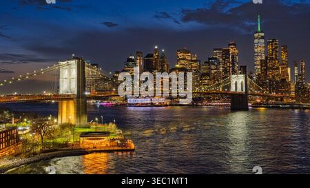 Die hellen Lichter von New York City erleuchten den Nachthimmel. Die Brooklyn Bridge überspannt das glühende Wasser, während im Hintergrund Wolkenkratzer aufsteigen. Stockfoto