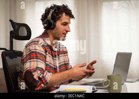 Junger Mann, der ein Notebook mit Headset nutzt, an Online-Kursen und virtuellen Meetings zu Hause teilnimmt. Stockfoto