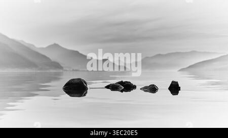 Schwarz-weiß minimalistischer Blick auf Felsen, die aus dem Comer See mit den Bergen in der Lombardei, Italien, auftauchen Stockfoto