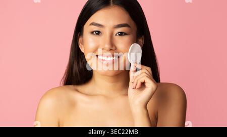 Die junge Frau lächelt, während sie ein Wattepad vor einem rosa Hintergrund hält Stockfoto