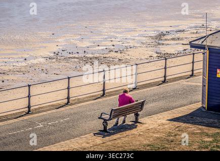 Eine Frau sitzt auf einer Bank an einer Strandpromenade. Vor ihr befindet sich ein Strand mit Wasser im Sand, der von einer rückläufigen Flut hinterlassen wurde. Ein Gebäude ist t Stockfoto