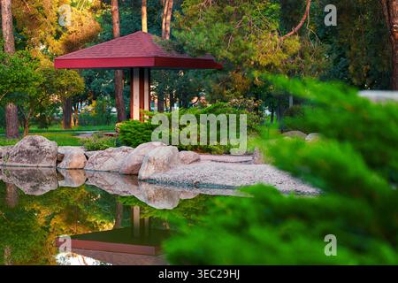 Goldenes Sonnenlicht taucht in eine ruhige Parkszene mit einem Pavillon im japanischen Stil, ruhigen, vom Wasser reflektierenden Bäumen und großen Felsen entlang des Randes auf Stockfoto