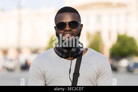 Stilvoller junger Mann mit Kopfhörern, der einen sonnigen Tag im Freien in einer Stadt genießt Stockfoto