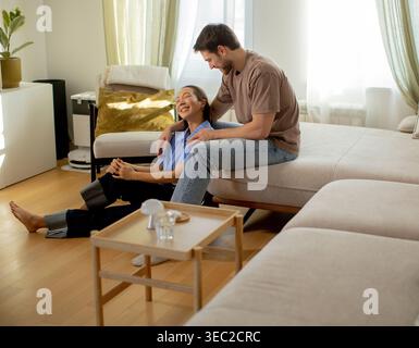 Eine Koreanerin sitzt lächelnd auf dem Boden, während ein Kaukasier auf einer Couch in der Nähe sitzt. Sie sind in einem hellen Raum mit sanftem Sonnenlicht und Holzboden Stockfoto