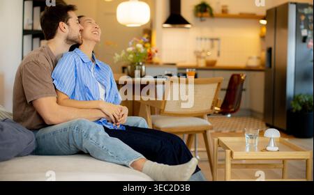 Eine Koreanerin sitzt mit einem Kaukasier auf einem Sofa. Sie genießen die Gesellschaft des anderen in einem lichtdurchfluteten Wohnzimmer. Blumen stehen auf einem Tisch, Stockfoto