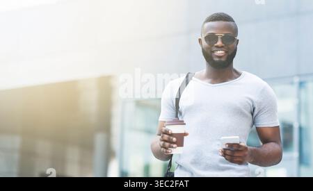Ein lächelnder Mann genießt tagsüber Kaffee und Smartphone in urbaner Umgebung Stockfoto