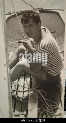 NOVI SAD, JUGOSLAWIEN (HEUTE SERBIEN) – 1968: Ein klassisches Schwarz-weiß-Foto eines jungen Mannes, Mitglied des Segelclubs „Vojvodina“ (Jedriličarski klub Vojvodina), der sich auf die Segel auf der Donau vorbereitet. Er sitzt in einem Rennboot der Finn-Klasse, trägt ein klassisches gestreiftes nautisches Hemd und hält die Takelage. Das Bild fängt die Sportkultur und die Traditionen des Freizeitsegels in Novi Sad in den späten 1960er Jahren ein Stockfoto