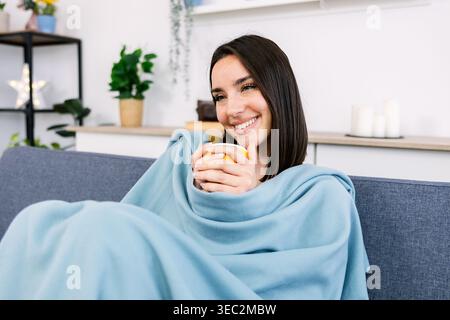 Lächelnde Frau in Decke gewickelt und genießt heißes Getränk auf dem Sofa Stockfoto