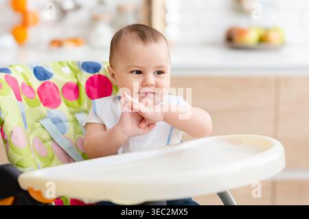 Happy Baby sitzt in einem Hochstuhl mit farbenfrohem Design in einer hellen Küche Stockfoto