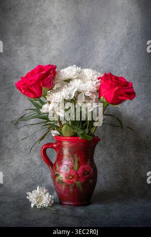 Stillleben von roten Rosen und weißen Chrysanthemen in einer rustikalen roten Keramikvase vor strukturiertem grauen Hintergrund. Stockfoto