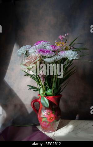 Elegantes Stillleben mit einer Mischung aus rosafarbenen Chrysanthemen und einer hellen Rose in einem roten Keramikkrug, hervorgehoben durch dramatisches Sonnenlicht und Schatten Stockfoto