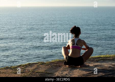 Eine Frau sitzt in einer Yoga-Pose auf einem felsigen Felsvorsprung mit Blick auf den weiten Ozean. Die Sonne geht unter, wirft warme Farben über den Himmel und verstärkt das ruhige Atmosp Stockfoto