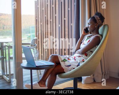Breites Seitenporträt einer jungen Frau, die in einem Schlafzimmer multitaskt, während sie telefoniert, während sie nach draußen blickt. Stockfoto