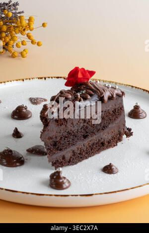Ein Stück reichhaltiger Schokoladenkuchen mit roter Blume, serviert auf einem weißen Teller mit Schokoladentropfen und Puderzucker-Dekoration Stockfoto