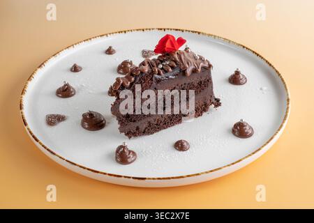 Ein Stück reichhaltiger Schokoladenkuchen mit roter Blume, serviert auf einem weißen Teller mit Schokoladentropfen und Puderzucker-Dekoration Stockfoto