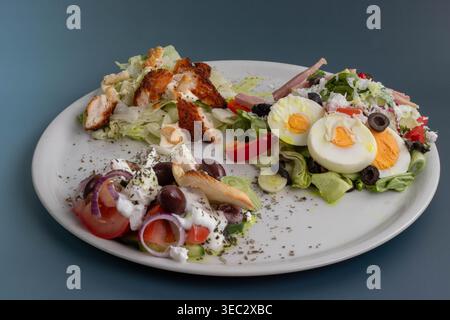 Blick von oben auf einen weißen Teller mit drei frischen Salaten: Knuspriges Chicken Caesar, griechischer Salat und Kochsalat mit gekochten Eiern. Stockfoto