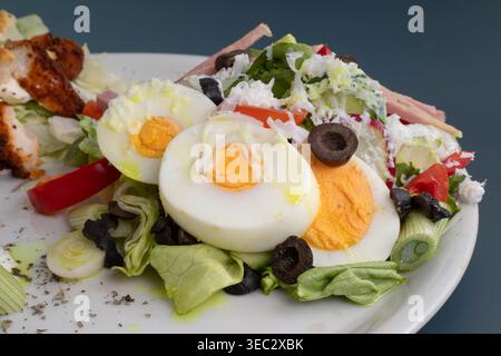 Nahaufnahme eines frischen Chefsalats mit gekochten Eierscheiben, schwarzen Oliven, geschnittenen grünen Zwiebeln und Schinken auf einem weißen Teller. Stockfoto