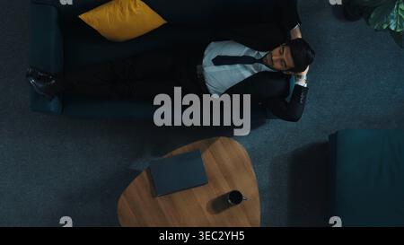 Von oben nach unten auf einem Laptop arbeitender Geschäftsmann, während er auf dem Sofa lag, um Stress zu lindern. Stressiger Mitarbeiter, der über einen Geschäftsplan oder eine Strategie nachdenkt Stockfoto