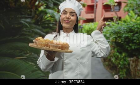 Die Chefin in weißem Mantel hält mit einer Hand ein hölzernes Muffins-Tablett und zeigt mit dem Finger auf den Weg zum Bau des Hofes; Backfreude. Stockfoto