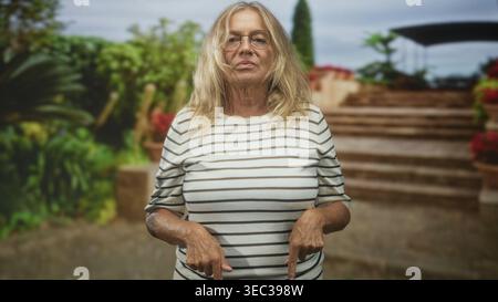 Eine ältere Frau zeigt mit den Zeigefingern auf eine Steintreppe im Gebäude, trägt ein gestreiftes Hemd und eine Brille; trotzt dem Stoizismus. Stockfoto