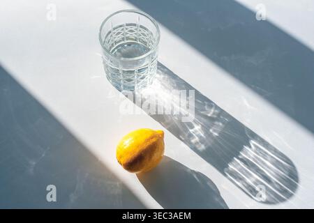 Minimales Stillleben mit einem Glas Wasser und frischer Zitrone, die im Sonnenlicht scharfe Schatten auf dem weißen Tisch werfen. Draufsicht. Stockfoto