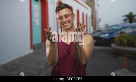 Junger blonder Mann, der mit beiden Händen Fingerherz macht und auf einer bunten Straße bei einem weißen Bogengebäude lächelt; verspielte Zuneigung. Stockfoto