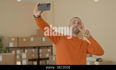Ein Mann im orangefarbenen Pullover hält das Smartphone für Selfie und zeigt mit der Hand ein Friedenszeichen zwischen gestapelten Kartonschachteln in einem Gebäude; Stolz auf Kleinunternehmen. Stockfoto