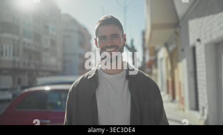 Junger hispanischer Mann lächelt mit sichtbarem Gesicht und Bart, trägt weißes T-Shirt und offenes Hemd, während er auf einer sonnigen Straße steht; Optimismus ist erreichbar Stockfoto