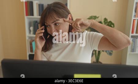 Frau, die auf dem Smartphone spricht, zeigt mit dem Finger auf den Tempel im Büro, wobei der Computerbildschirm sichtbar ist; nachdenklicher Fokus. Stockfoto