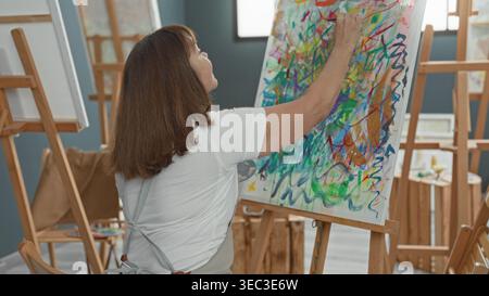 Malerin steht an der Staffelei, rechte Hand malt dicke bunte Striche auf Leinwand, während sie Palette hält, Rückansicht im Kunststudio; Konzentration cr Stockfoto