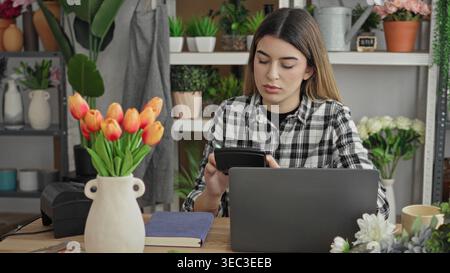 Floristin mit Taschenrechner und Laptop, Hand bis Stirn im Blumenstudio; finanzieller Stress im Fokus. Stockfoto