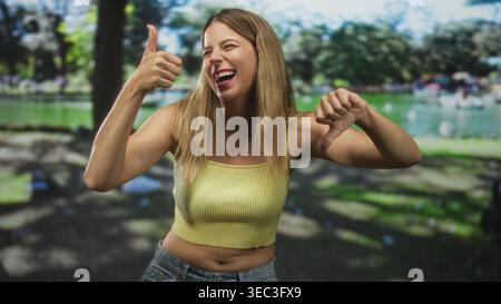 Frau in einem gelben, abgeschnittenen Tanktop zeigt die Daumen hoch und Daumen nach unten Gesten mit ausdrucksstarkem Gesicht in einem Studio-Set, das einem Park ähnelt; Unentschlossenheit Zweifel. Stockfoto